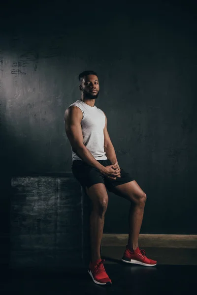 Full length view of young athletic american man in sportswear sitting and looking at camera on black — Fotografia de Stock