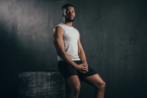 Joven atlético afroamericano hombre en ropa deportiva sentado y mirando a la cámara en negro - foto de stock