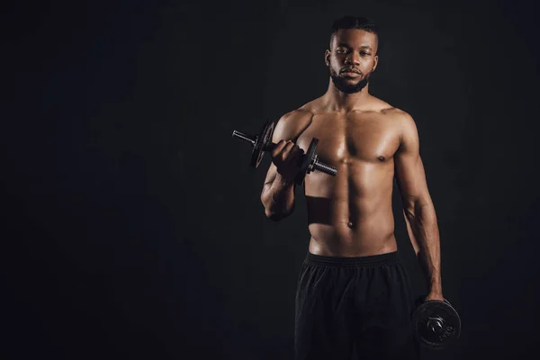 Musculoso sin camisa afroamericano deportista haciendo ejercicio con mancuernas y mirando a la cámara aislada en negro - foto de stock
