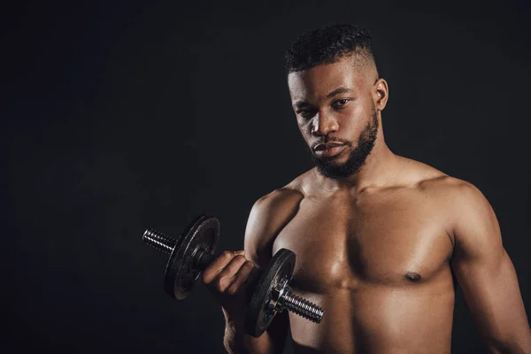 Musculoso sin camisa afroamericano deportista entrenamiento con mancuerna y mirando a la cámara aislado en negro - foto de stock