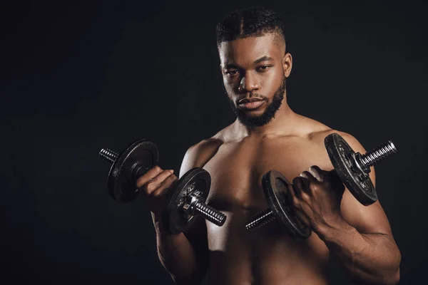 Muskulöser, hemdsloser afrikanisch-amerikanischer Sportler, der mit Kurzhanteln trainiert und isoliert auf schwarz in die Kamera schaut — Stockfoto
