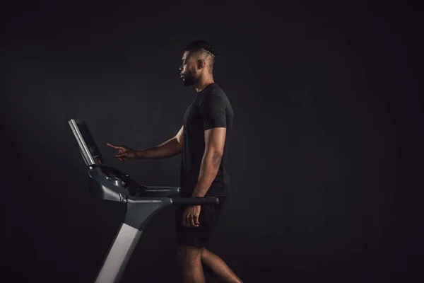 Vue latérale du jeune homme athlétique afro-américain faisant de l'exercice sur tapis roulant isolé sur noir — Photo de stock