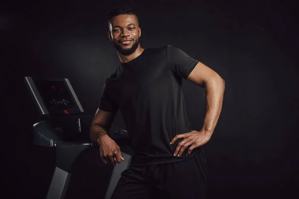 Handsome young athletic african american man leaning at treadmill and smiling at camera — Stock Photo