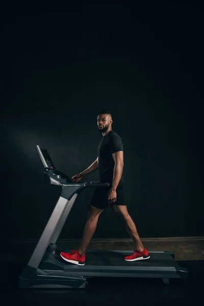 Joven deportista afroamericano ejercitándose en la cinta de correr y mirando la cámara en negro — Stock Photo