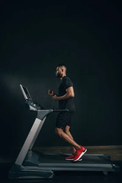 Visão lateral do jovem esportista afro-americano correndo na esteira em preto — Fotografia de Stock