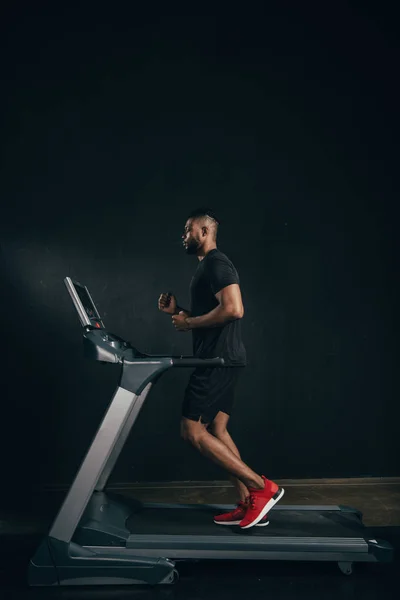 Vue latérale du jeune sportif afro-américain courant sur tapis roulant sur noir — Photo de stock