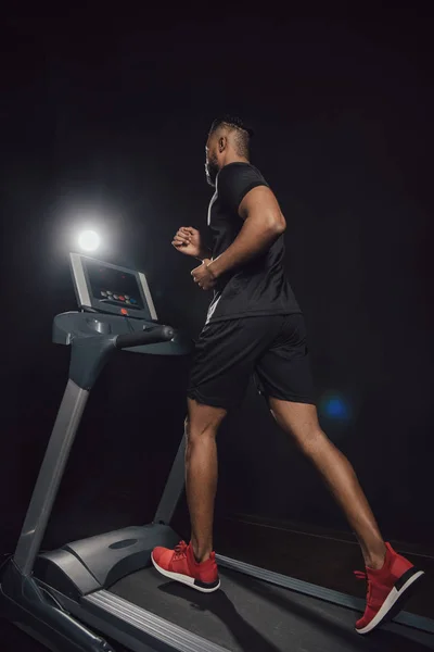 Low angle view of young african american sportsman jogging on treadmill on black — Stock Photo