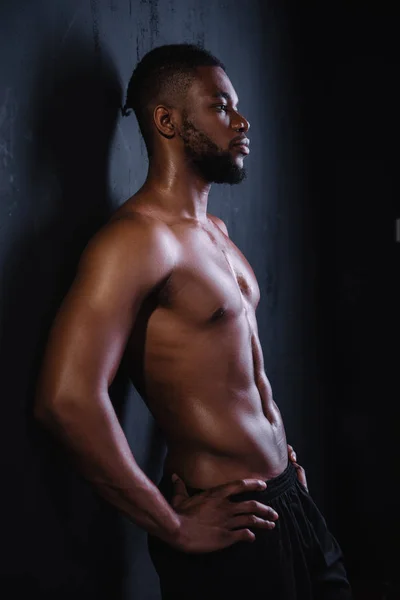 Vista lateral del joven afroamericano musculoso sin camisa de pie con las manos en la cintura y mirando hacia otro lado en negro - foto de stock