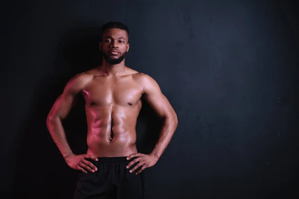 Musculoso hombre afroamericano con el pecho desnudo de pie con las manos en la cintura y mirando a la cámara aislada en negro - foto de stock