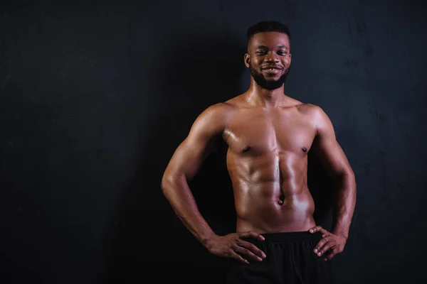 Sin camisa musculoso joven afroamericano hombre de pie con las manos en la cintura y sonriendo a la cámara en negro - foto de stock