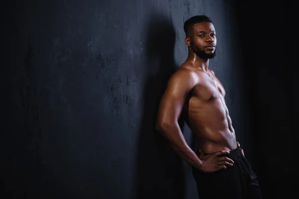 Sin camisa musculoso joven afroamericano hombre de pie con las manos en la cintura y mirando a la cámara en negro - foto de stock