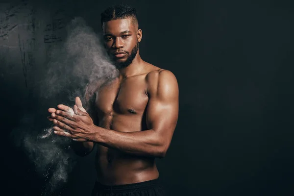 Joven desnudo-pecho afroamericano deportista aplicando talco en polvo y mirando a la cámara en negro - foto de stock