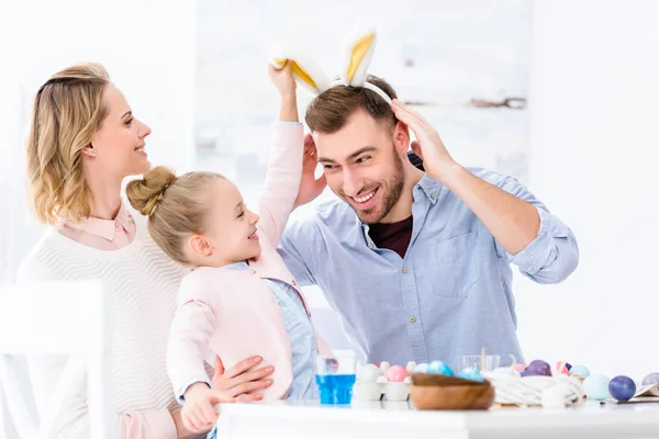 Smiling family having fun with bunny ears by table with painted eggs on Easter — Stock Photo