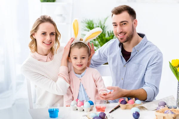 Celebrando familia con hija jugando con orejas de conejo de Pascua por mesa con huevos pintados - foto de stock