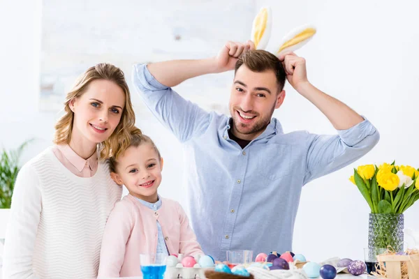 Familia feliz jugando con orejas de conejo por mesa con huevos de colores para Pascua - foto de stock