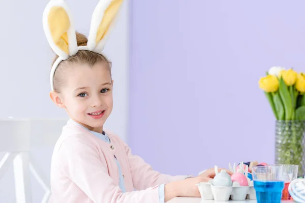 Menina criança jogando vestindo orelhas de coelho por mesa com ovos de cor de Páscoa — Fotografia de Stock