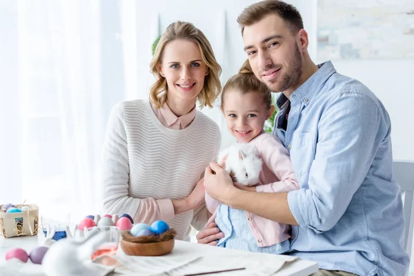 Familia feliz sosteniendo conejo de Pascua con huevos de Pascua en la mesa - foto de stock
