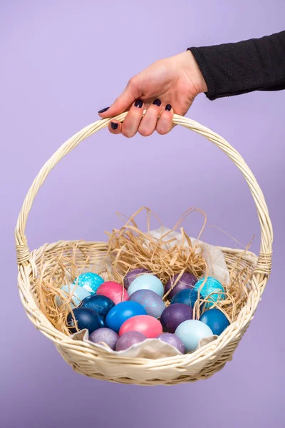 Female hand with Easter basket with colored eggs isolated on violet — Stock Photo