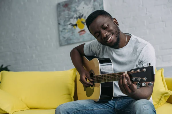 Guapo sonriente joven afroamericano hombre tocando la guitarra en casa - foto de stock