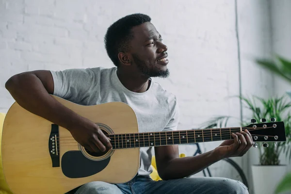 Sonriente joven afroamericano hombre tocando la guitarra acústica y mirando hacia otro lado - foto de stock