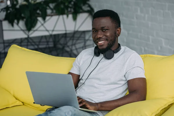 Young african american man in headphones sitting on sofa with laptop and smiling at camera — Stock Photo