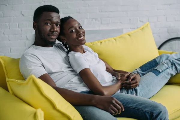 Heureux jeune couple afro-américain assis sur canapé et souriant à la caméra — Photo de stock