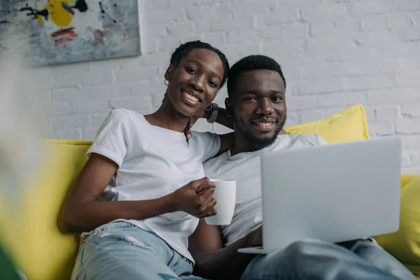 Feliz jovem casal afro-americano com laptop e xícara de chá sorrindo para a câmera — Fotografia de Stock