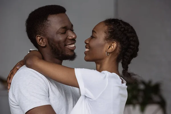 Feliz jovem casal afro-americano abraçando e sorrindo uns aos outros em casa — Fotografia de Stock