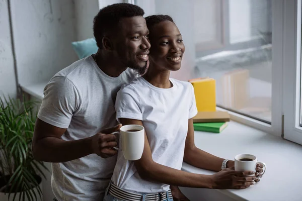 Happy young african american couple holding cups of coffee and looking at window — Stock Photo