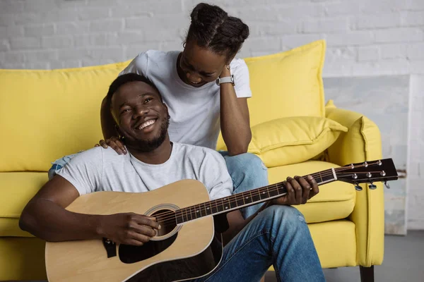 Jeune femme afro-américaine assise sur le canapé tandis que son petit ami heureux joue de la guitare — Photo de stock