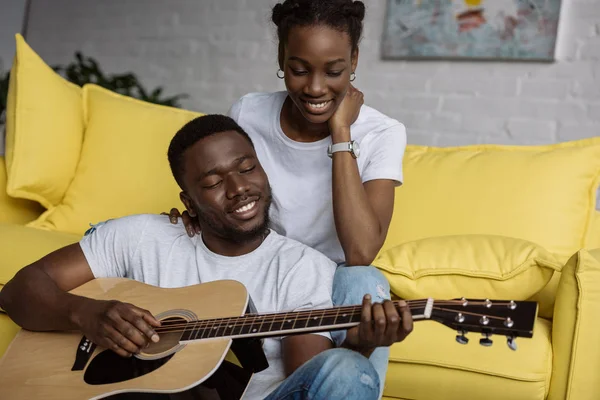 Glückliche junge afrikanisch-amerikanische Frau sieht ihren Freund zu Hause Gitarre spielen — Stockfoto