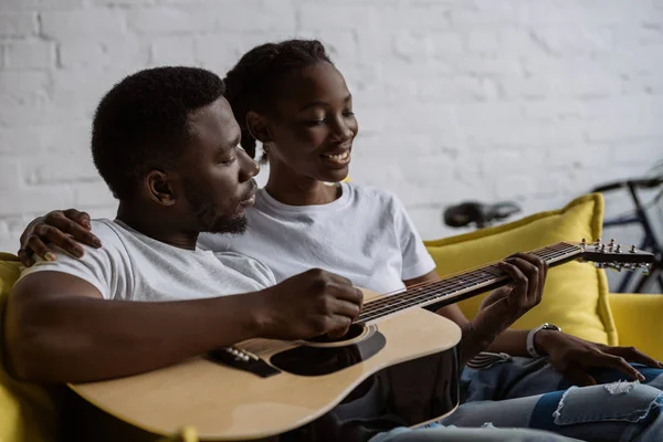 Щаслива молода афроамериканська пара сидить разом на дивані і чоловік грає на акустичній гітарі — стокове фото