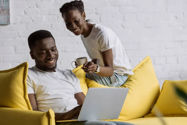 Sourire jeune couple afro-américain en utilisant un ordinateur portable tout en étant assis sur le canapé à la maison — Photo de stock