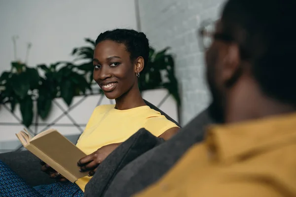 Foyer sélectif de belle jeune femme afro-américaine souriante tenant livre et regardant petit ami à la maison — Photo de stock