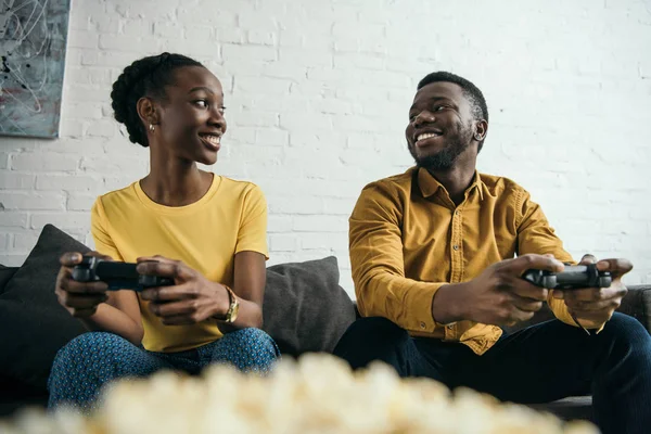 Glückliches junges afrikanisch-amerikanisches Paar, das mit Joysticks spielt und sich gegenseitig ansieht — Stockfoto