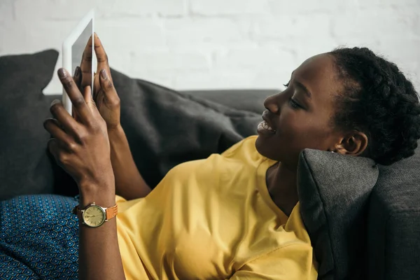Smiling young african american woman using digital tablet while lying on sofa at home — Stock Photo