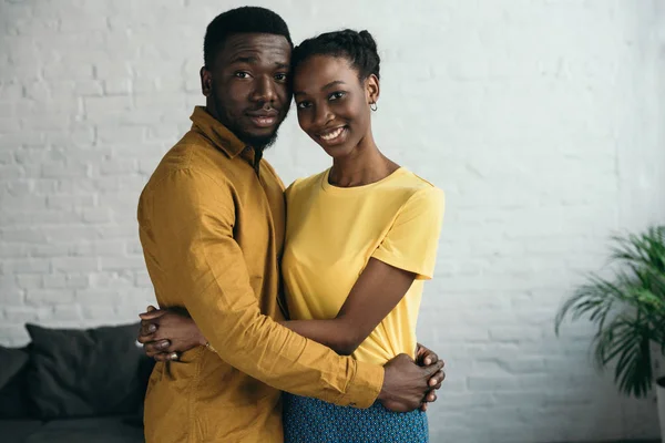 Красивая счастливая молодая африканская пара, обнимающая и улыбающаяся перед камерой дома — стоковое фото