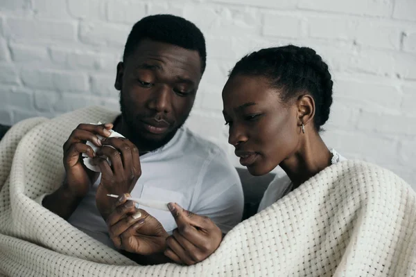 Молодая африканская американская пара проверяет термометр, пока они вместе болеют гриппом дома — стоковое фото