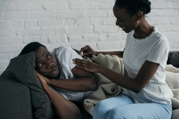 Souriant jeune femme afro-américaine vérifier la température du copain malade couché sur le canapé — Photo de stock