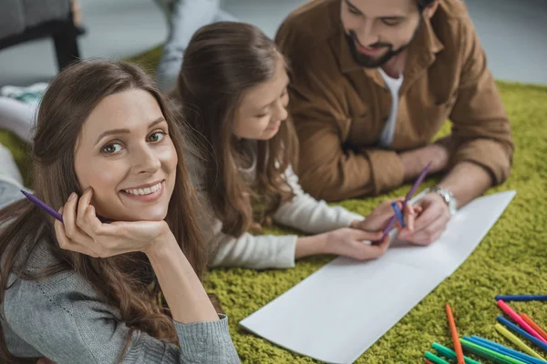 Sonriente esposa mirando a la cámara mientras hija y marido dibujando con rotuladores - foto de stock