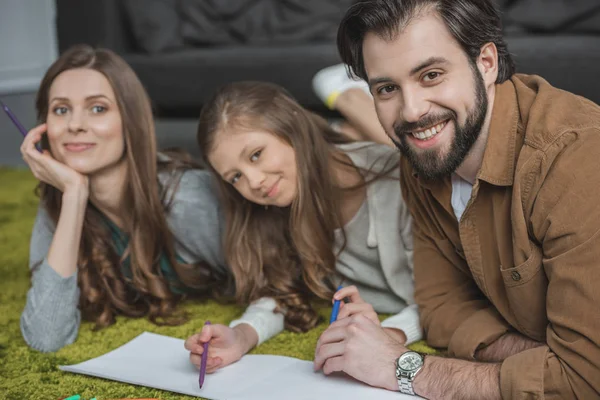 Happy parents and daughter drawing with felt-tip pens on floor and looking at camera — Stock Photo