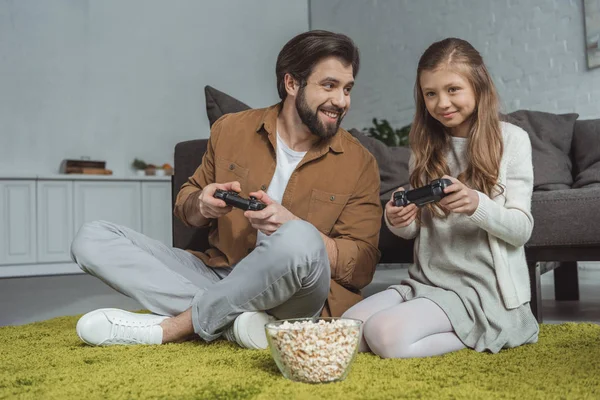 Padre e hija sentados en la alfombra y jugando videojuegos - foto de stock