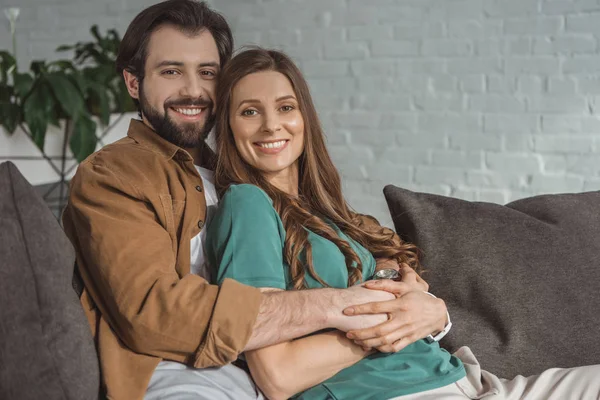 Novio sonriente abrazando novia y mirando a la cámara en casa - foto de stock