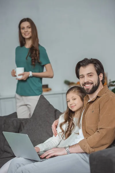 Padre e figlia sorridenti seduti con il computer portatile sul divano e guardando la fotocamera — Foto stock