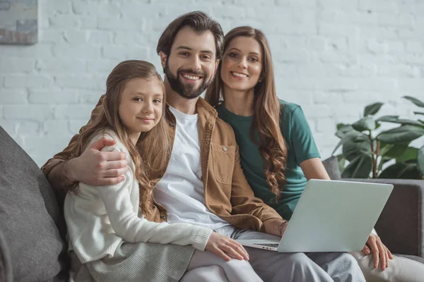 Glückliche Eltern und Tochter sitzen mit Laptop und blicken in die Kamera — Stockfoto