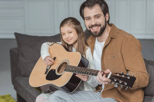 Улыбающийся отец учит дочь играть на акустической гитаре и смотреть в камеру — стоковое фото