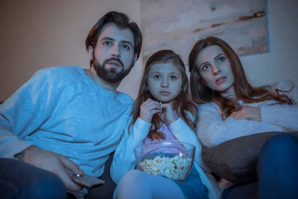 Padres sorprendidos y su hija viendo películas en el sofá con palomitas de maíz - foto de stock