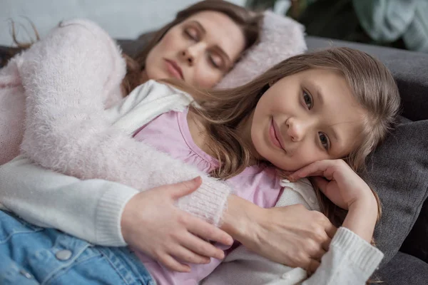 Mãe dormindo no sofá e filha olhando para a câmera em casa — Fotografia de Stock