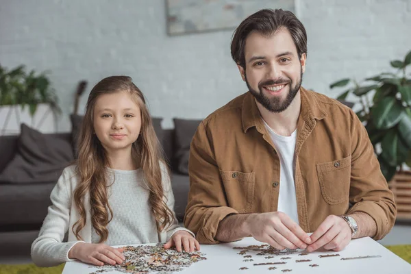 Happy father and daughter sitting at table with puzzle pieces and looking at camera — Stock Photo