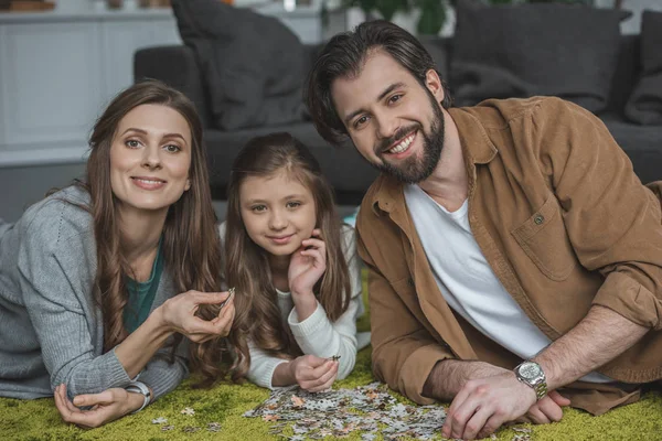 Lächelnde Eltern und Tochter liegen mit Puzzleteilen auf Teppich und schauen in die Kamera — Stockfoto